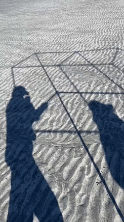 a gif of two people walking with portable bus stop you can see the shadow of the structure moving over the sand
